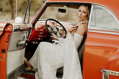 A bride sitting in the driver's seat of a red vintage car, representing the unique options for wedding transportation in Fredericksburg TX