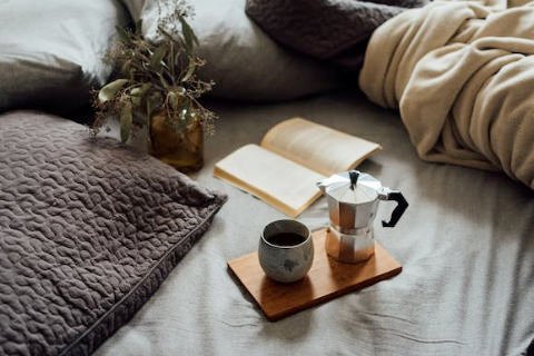 A closeup of a bed with a coffee pot and mug, an open book, and plant on it, representing the bed and breakfasts and other places to stay in Fredericksburg TX