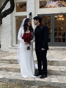A bride and groom smiling and looking at each other. The bride is holding a wedding bouquet of red roses