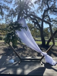 A wooden hexagon arch with white fabric draping and floral spray for a wedding in Fredericksburg TX, provided by A Better Fete