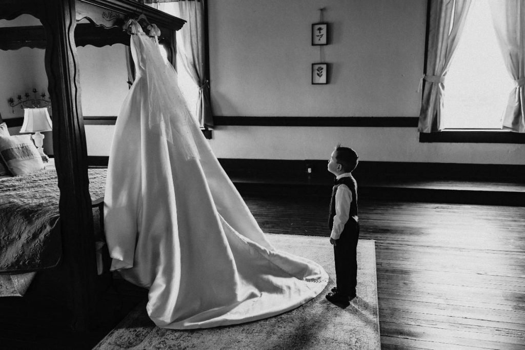 A black and white image of a ring bearer looking at the bride's wedding dress. Image captured by Kayla Jane Photography, a wedding photographer in Fredericksburg TX