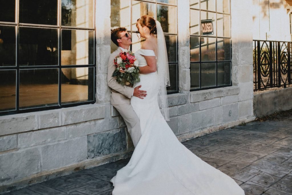 A bride and groom leaning against an exterior window smiling at each other. Image captured by Kayla Jane Photography, a wedding photographer in Fredericksburg TX