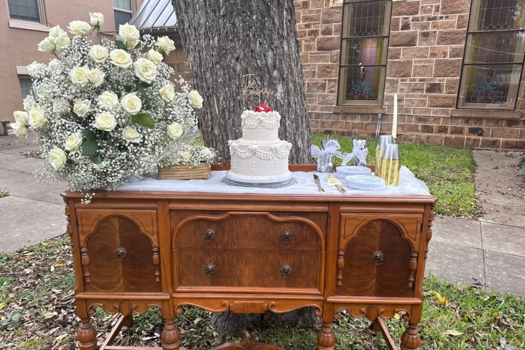 A small 2-tier cake outdoors on an antique dresser surrounded by flowers and cutlery. The cake was created by ROC Cakes and Cupcakes for weddings in Fredericksburg TX.
