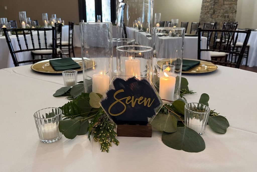 A wedding reception centerpiece with a black and gold table number, candles, and greenery