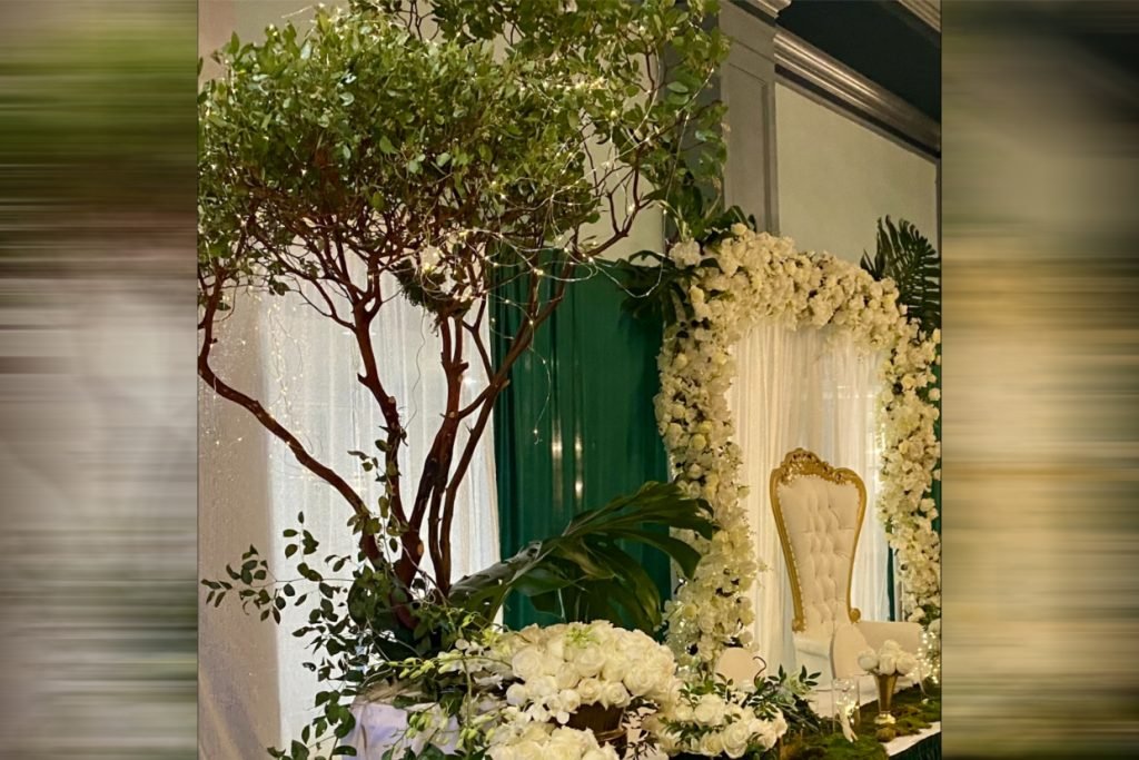 A tree with twinkle lights next to a whtie wedding floral arch. A gold and white throne sits under it.