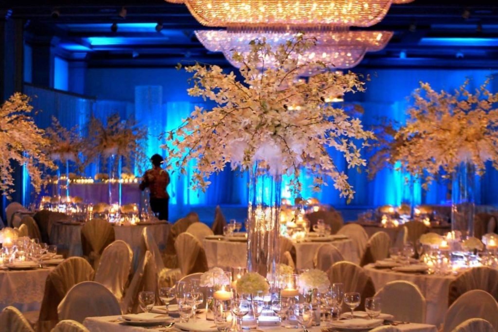 A group of wedding reception tables with tall wedding floral centerpieces. Candles and blue uplights light up the room.