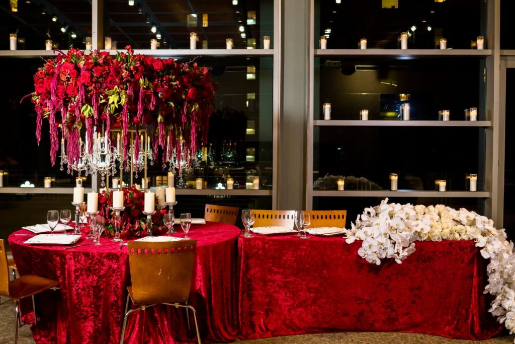 Two wedding reception tables with red velvet linens. A lavish red wedding floral centerpiece is on one table and a white floral garland is on the other.