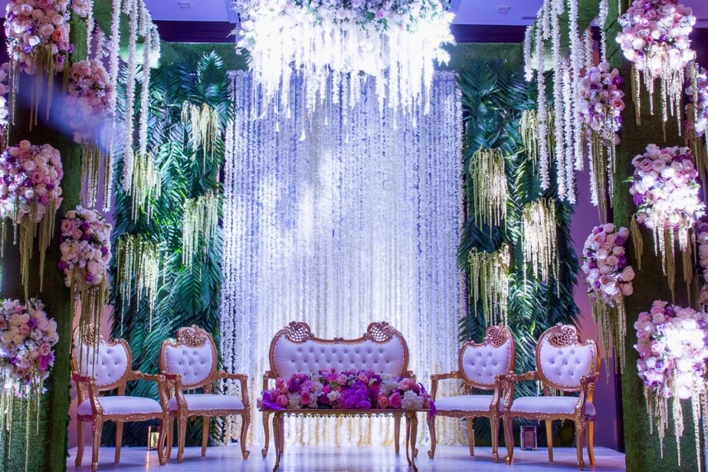 An Indian wedding mandap with a group of gold and white ornate chairs. This is surrounded by dripping wedding floral in pink, purple, white, and green, all provided by Bisli Luxurious Events