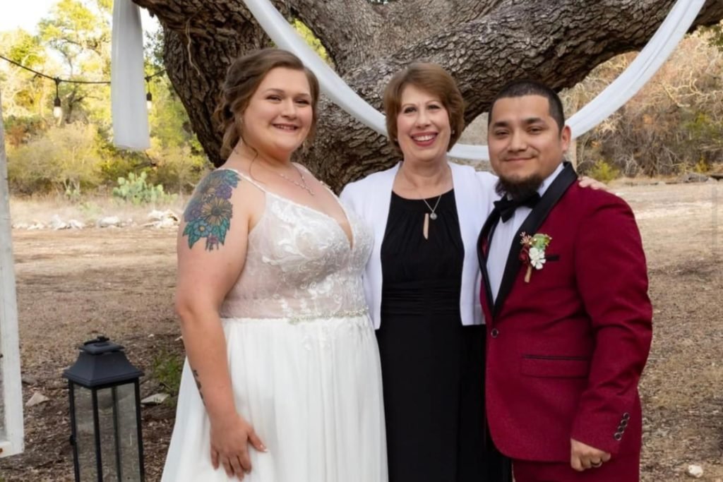 A female wedding officiant in Fredericksburg TX smiles and stands in between two newlyweds.
