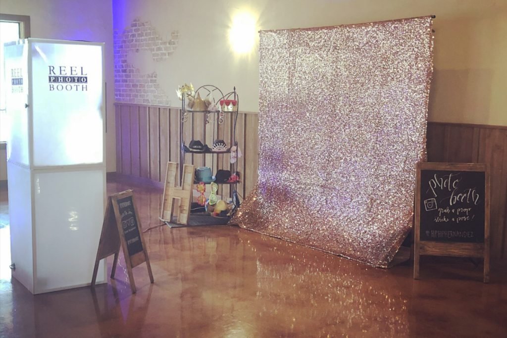 A gold glitter backdrop with a stand of props next to it, as part of a wedding photo booth provided by Reel Photo Booth